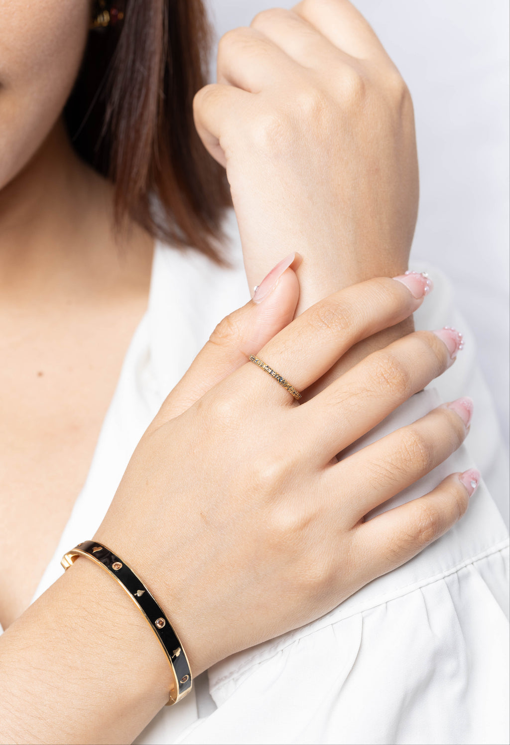 A close-up of a woman's hands wearing a black bangle bracelet with gold studs on one wrist and a thin gold name ring on the other hand, against a clean white background.