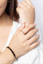 A close-up of a woman's hands wearing a black bangle bracelet with gold studs on one wrist and a thin gold name ring on the other hand, against a clean white background.