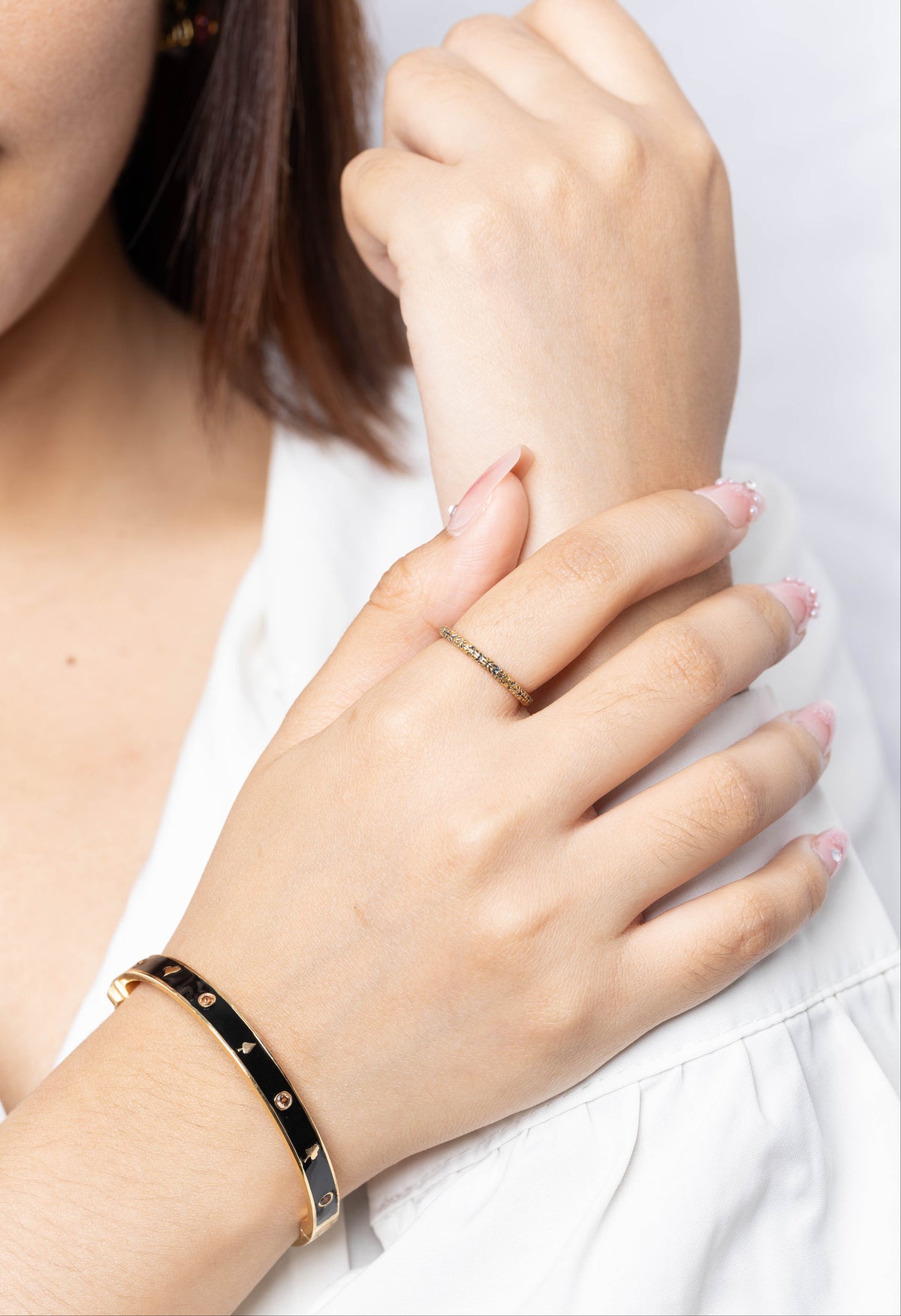A close-up of a woman's hands wearing a black bangle bracelet with gold studs on one wrist and a thin gold name ring on the other hand, against a clean white background.