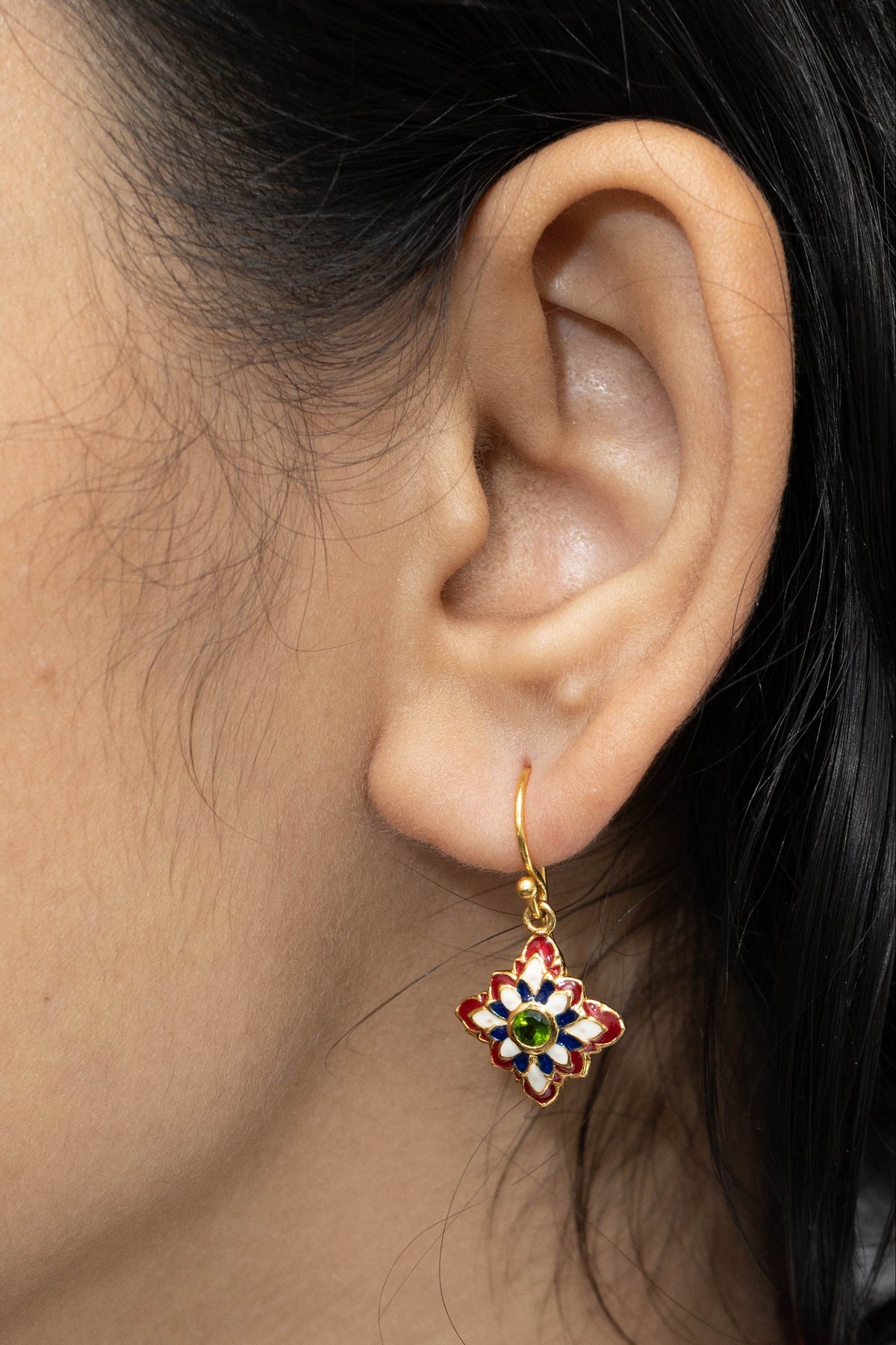 A close-up of a person's ear wearing a small gold hoop earring with a dangling four-petal flower charm decorated with red, green, blue, and white enamel.