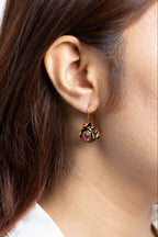A close-up of a woman's ear wearing a small dangling earring with a gold-tone hook and a decorative charm featuring black, red, and gold details.