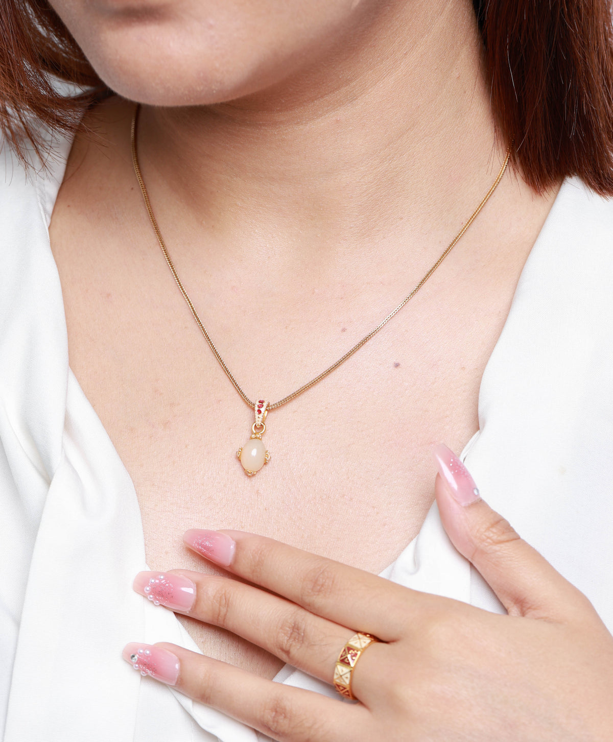 A close-up of a woman wearing a delicate gold necklace with a small heart-shaped pendant, a matching gold ring, and a white beaded bracelet, paired with a white blouse.