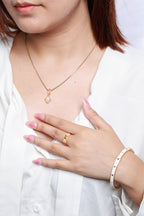 A close-up of a woman wearing a delicate gold necklace with a small heart-shaped pendant, a matching gold ring, and a white beaded bracelet, paired with a white blouse.