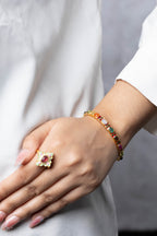 A close-up of a person's hand and wrist wearing a gold bracelet with colorful gemstones and a matching gold ring with a red stone, styled with a white top.