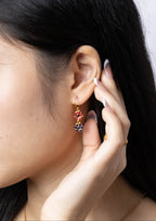 A close-up of a woman's ear wearing a gold dangle earring with colorful floral details, including red, blue, and yellow elements.
