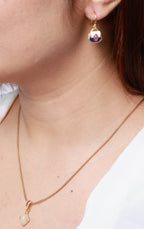A close-up lifestyle shot of a woman wearing a delicate gold chain necklace with a small pale stone pendant and a matching gold drop earring with a colorful, faceted gemstone.