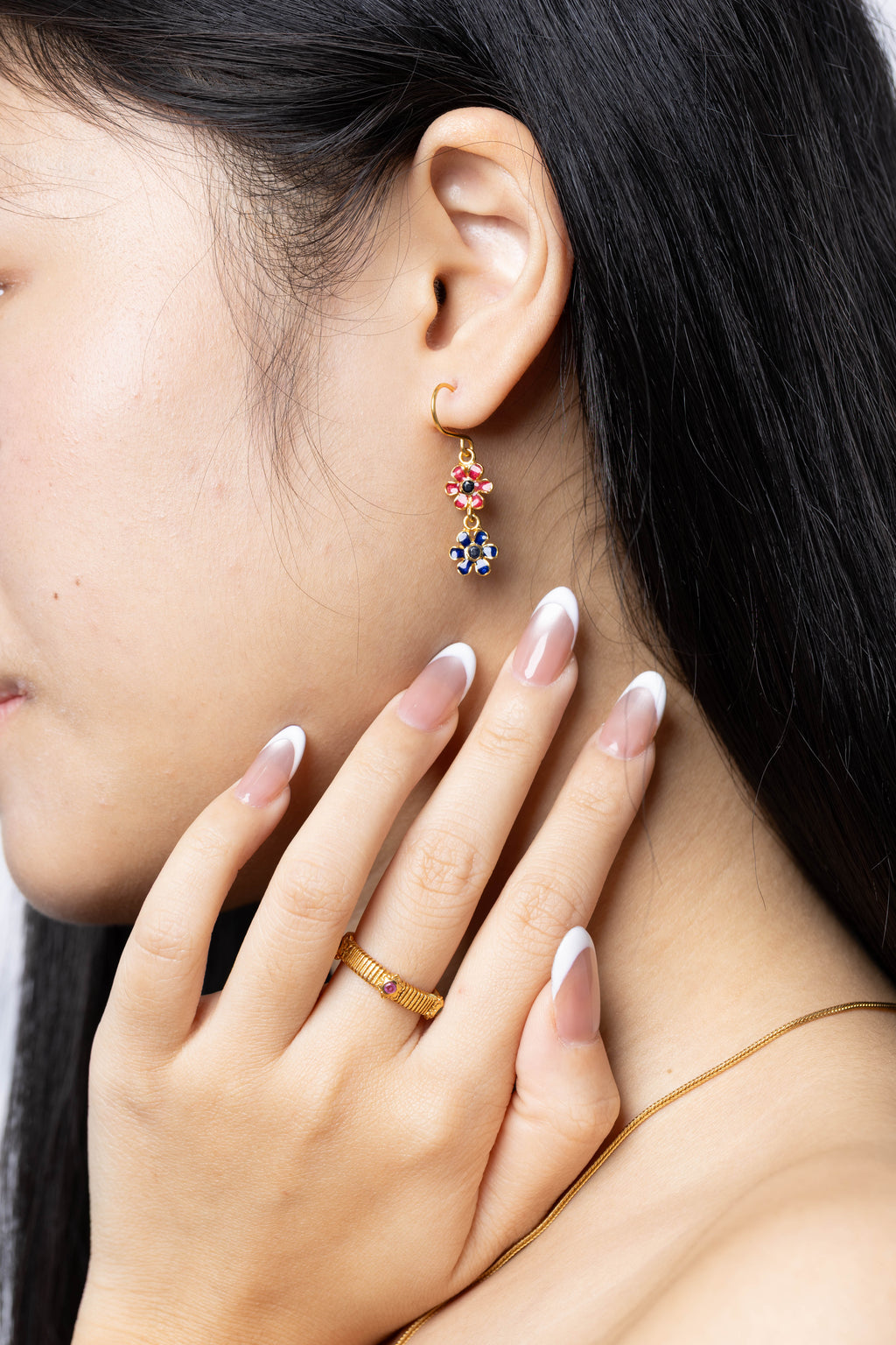 A close-up lifestyle shot of a model wearing a gold huggie hoop earring with a dangling floral charm featuring red and blue stones, paired with a gold ring and necklace.