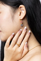 A close-up lifestyle shot of a model wearing a gold huggie hoop earring with a dangling floral charm featuring red and blue stones, paired with a gold ring and necklace.