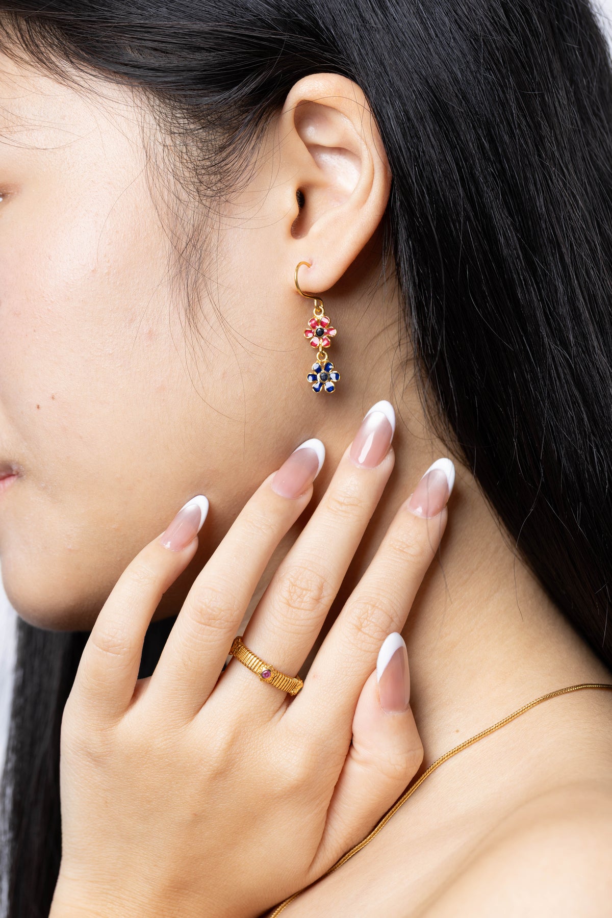 A close-up lifestyle shot of a model wearing a gold huggie hoop earring with a dangling floral charm featuring red and blue stones, paired with a gold ring and necklace.