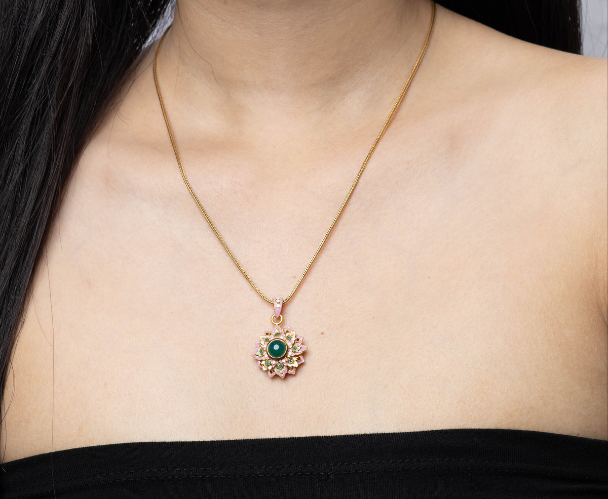A close-up of a woman wearing a delicate gold chain necklace with a circular pendant featuring a green center and pink surrounding stones, paired with a black strapless top.