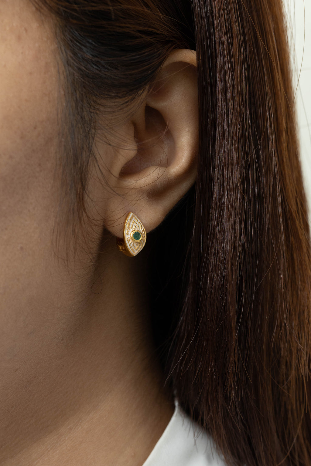 A close-up of a woman's ear wearing a small gold teardrop-shaped stud earring with a central dark green stone, against a light neutral background.