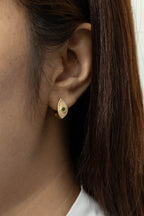 A close-up of a woman's ear wearing a small gold teardrop-shaped stud earring with a central dark green stone, against a light neutral background.