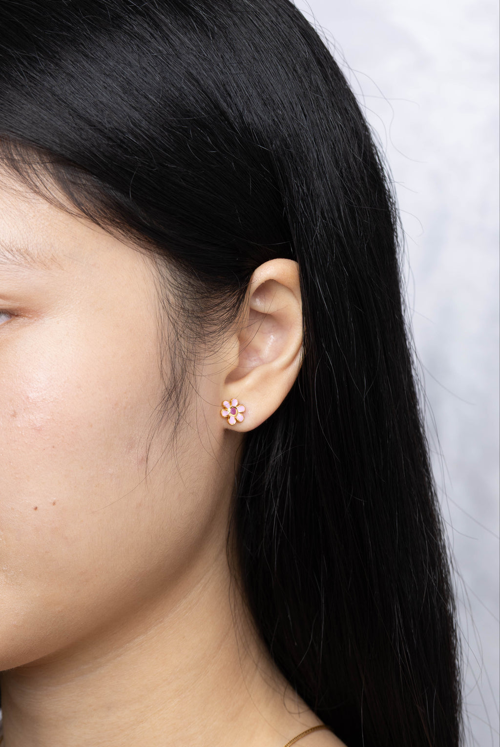 A close-up side profile of a woman with long black hair wearing a delicate gold chain necklace and a small gold stud earring with pink gemstones.