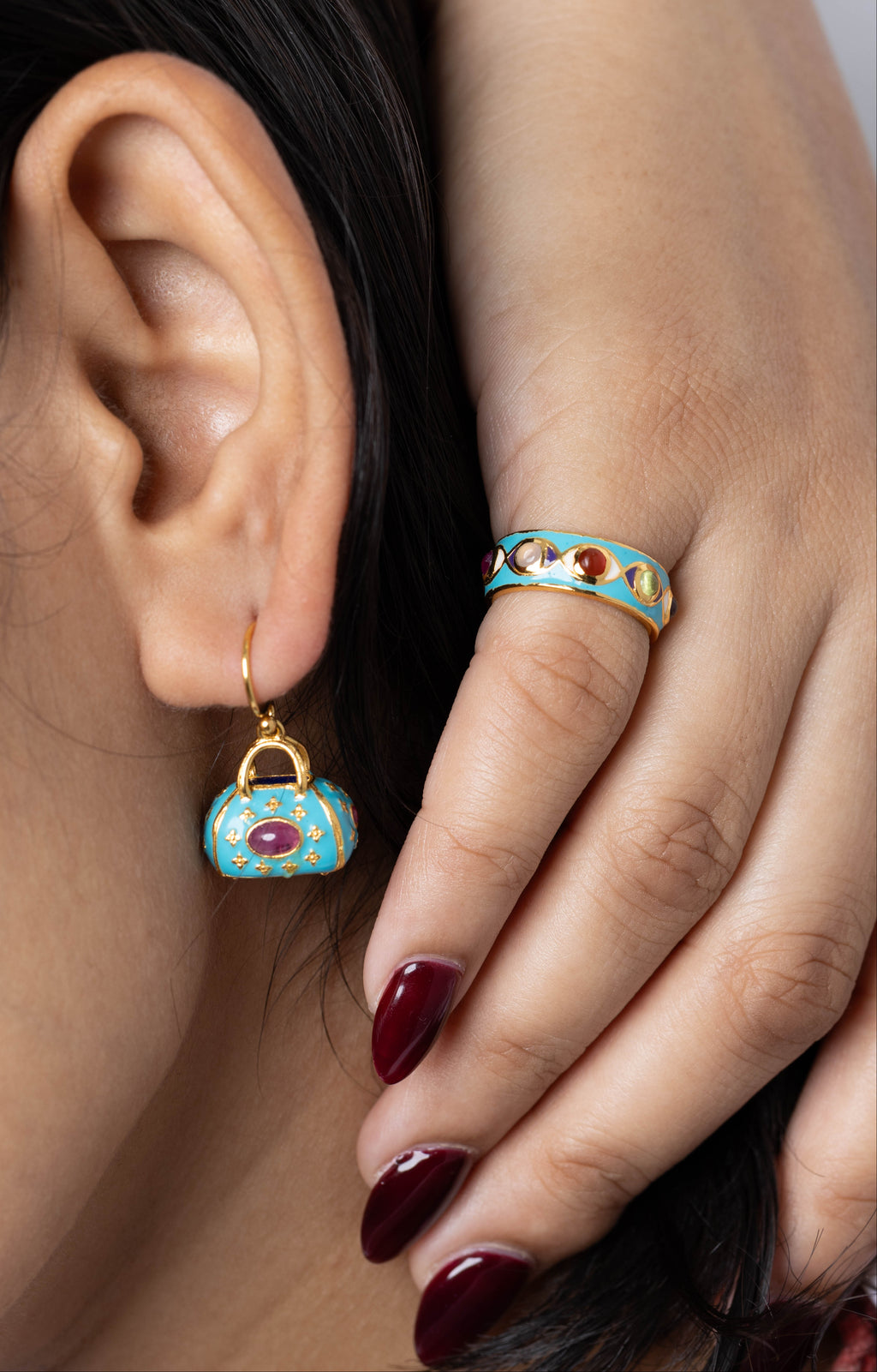 Close-up of a model wearing a small turquoise and gold handbag-shaped drop earring and a matching turquoise and gold ring with red gemstone accents.