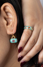 Close-up of a model wearing a small turquoise and gold handbag-shaped drop earring and a matching turquoise and gold ring with red gemstone accents.