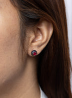 A close-up of a woman's ear wearing a small round stud earring with a dark blue outer ring and a red center stone.