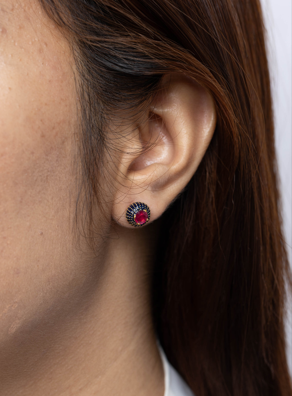 A close-up of a woman's ear wearing a small round stud earring with a dark blue outer ring and a red center stone.