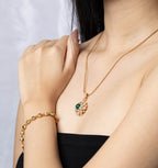 A close-up lifestyle shot of a model wearing a gold necklace with a floral pendant featuring a green gemstone and a matching gold bracelet.