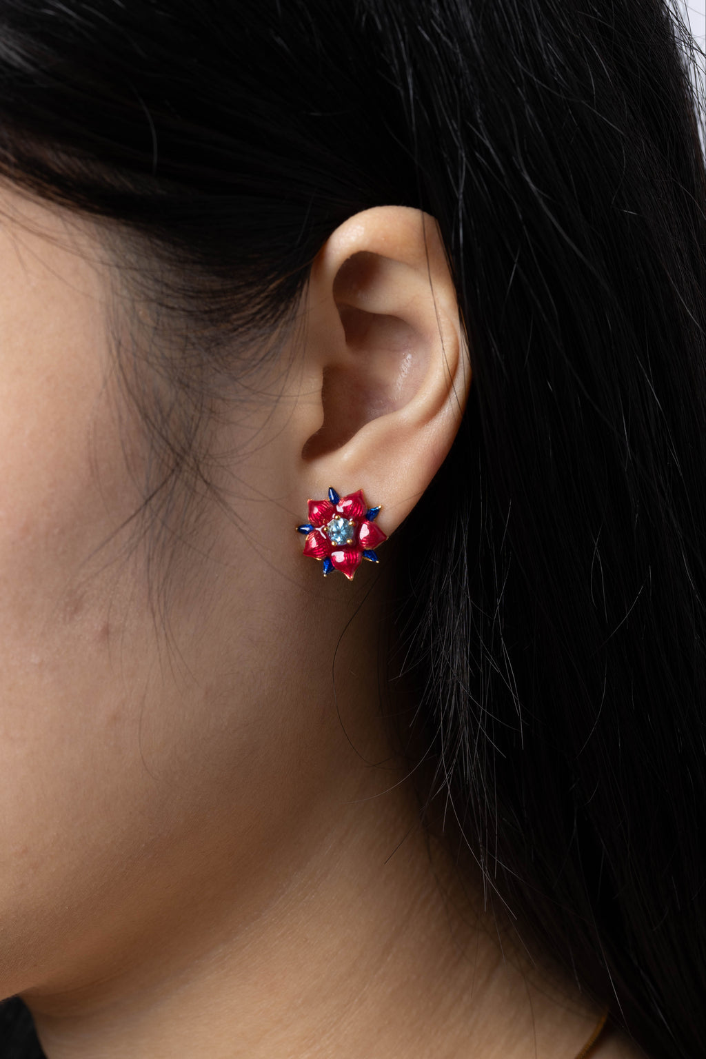 A close-up side profile of a woman wearing a small floral stud earring with red and pink stones and a gold-tone setting.