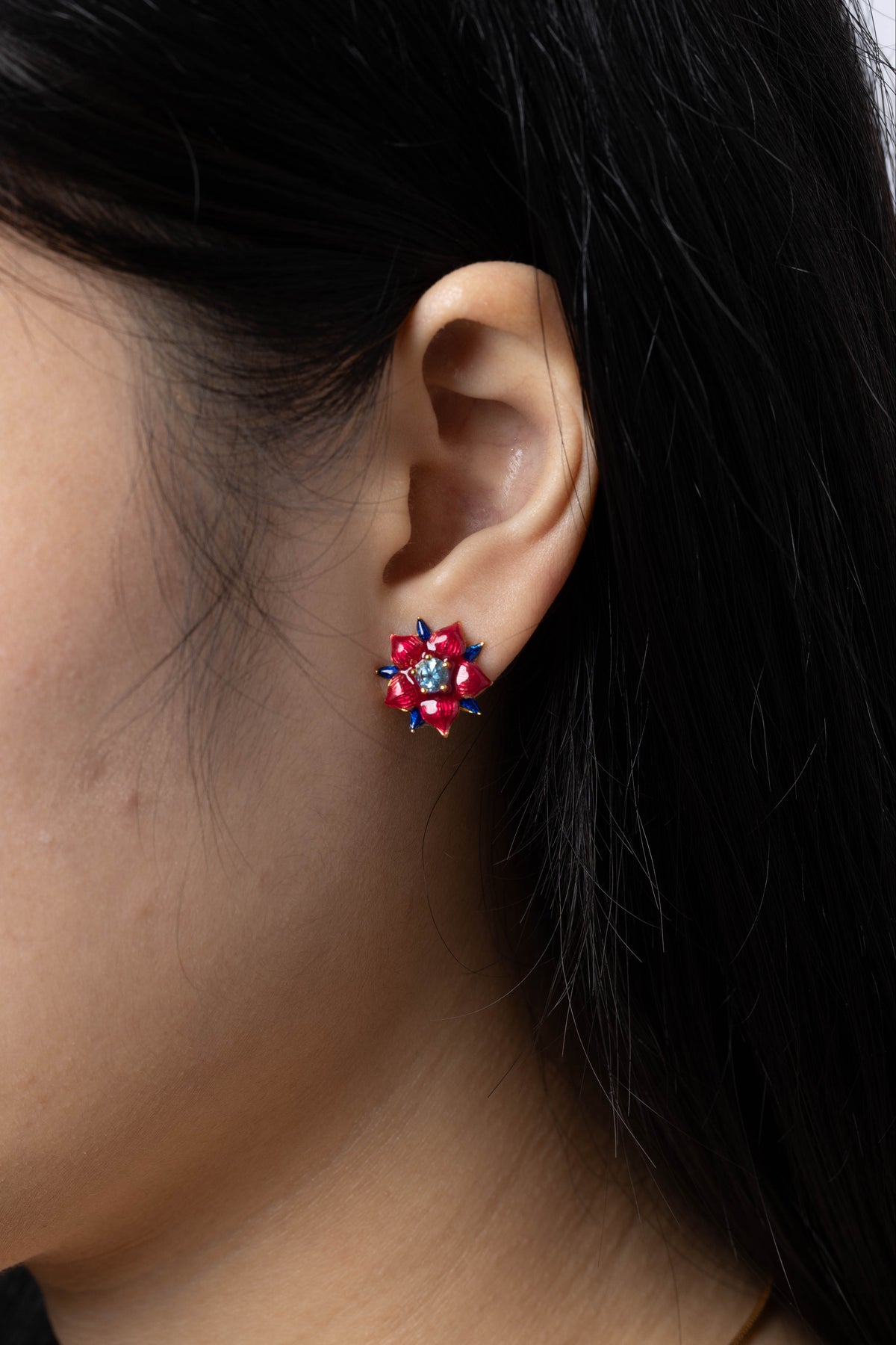 A close-up side profile of a woman wearing a small floral stud earring with red and pink stones and a gold-tone setting.