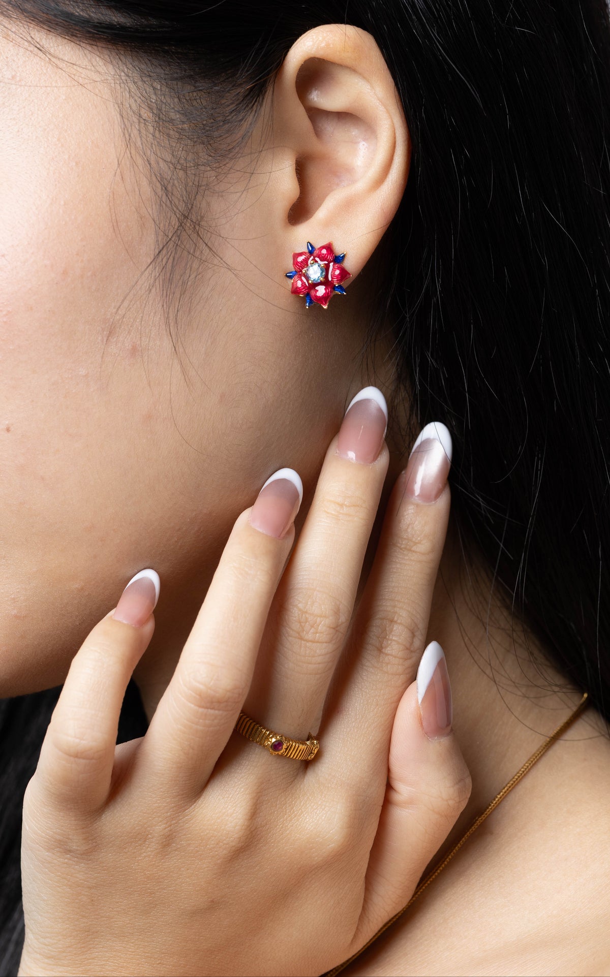 A close-up of a woman's ear wearing a small red and blue floral stud earring, with her hand resting near her neck. 