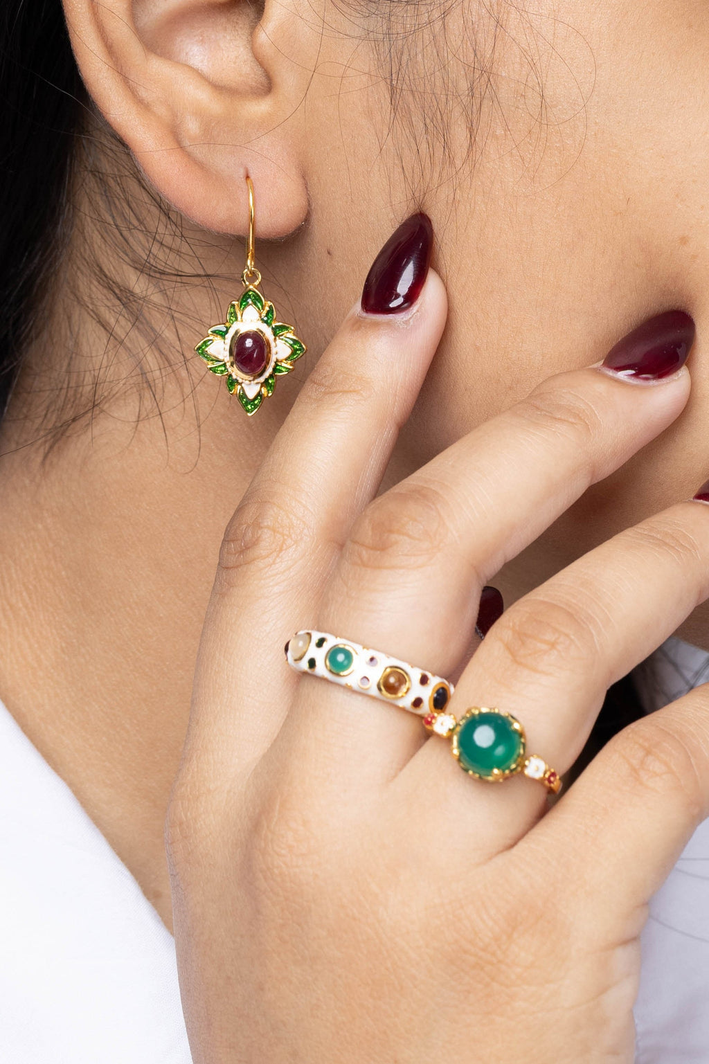 Close-up of a woman wearing a gold earring with a red and green gemstone and a matching gold ring with green and orange stones, touching her cheek.