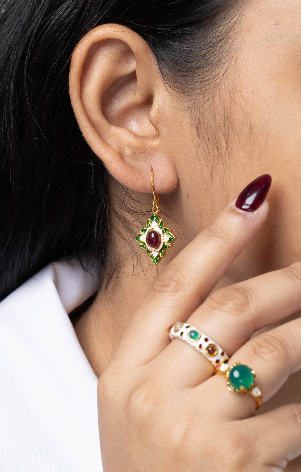 A close-up lifestyle shot of a woman wearing gold rings and earrings with green and red stones, touching her cheek. She is wearing a white collared shirt against a light gray background.