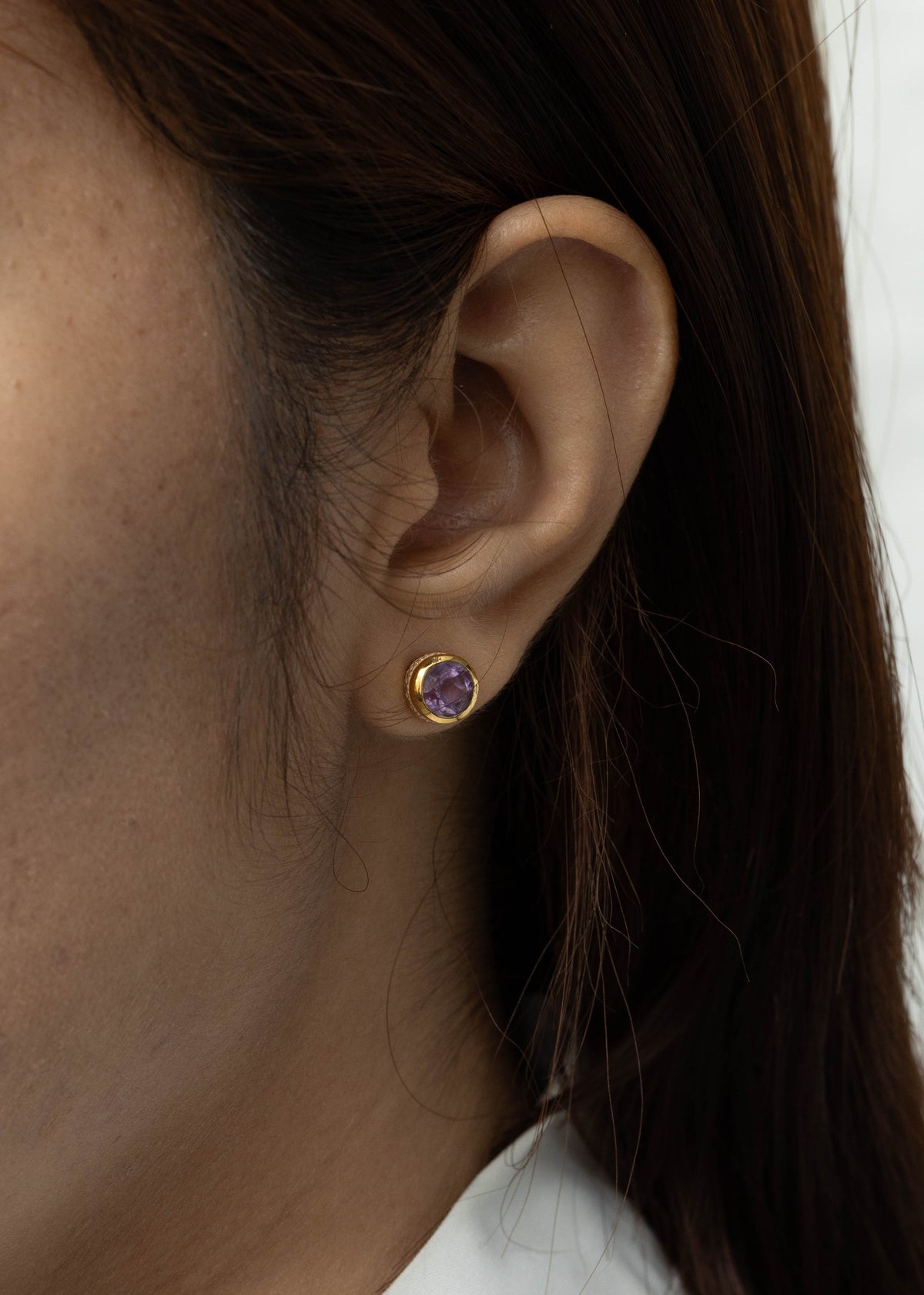 Close-up of a person's ear wearing a small gold stud earring with a purple gemstone, against a light neutral background.