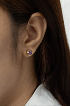 A close-up of a person's ear wearing a small gold stud earring with a round purple gemstone, against a light neutral background.