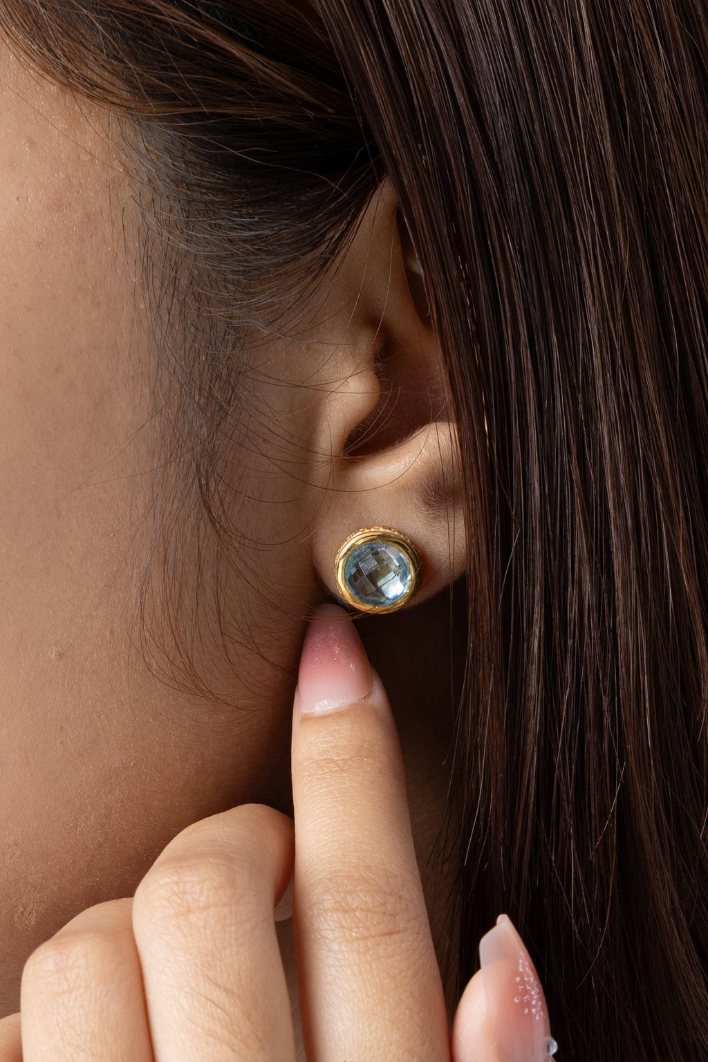 Close-up of a woman wearing a gold ring with a blue gemstone on her finger and a matching gold stud earring with a blue gemstone on her earlobe.