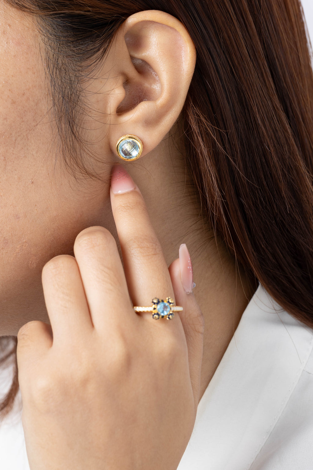Close-up of a woman wearing a small round stud earring with a light blue stone and gold setting, with her hand near her face. She is also wearing a gold ring with a blue stone.