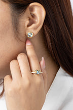 Close-up of a woman wearing a small round stud earring with a light blue stone and gold setting, with her hand near her face. She is also wearing a gold ring with a blue stone.
