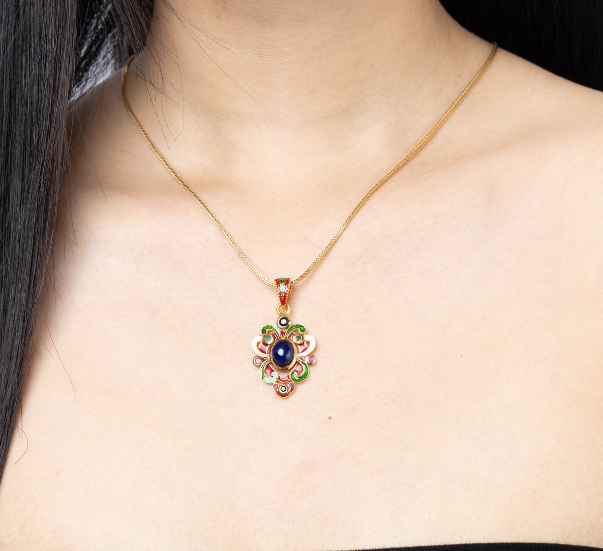 A woman with long black hair wearing a gold chain necklace with a colorful pendant featuring red, blue, and green stones, paired with a black strapless top against a light gray background.