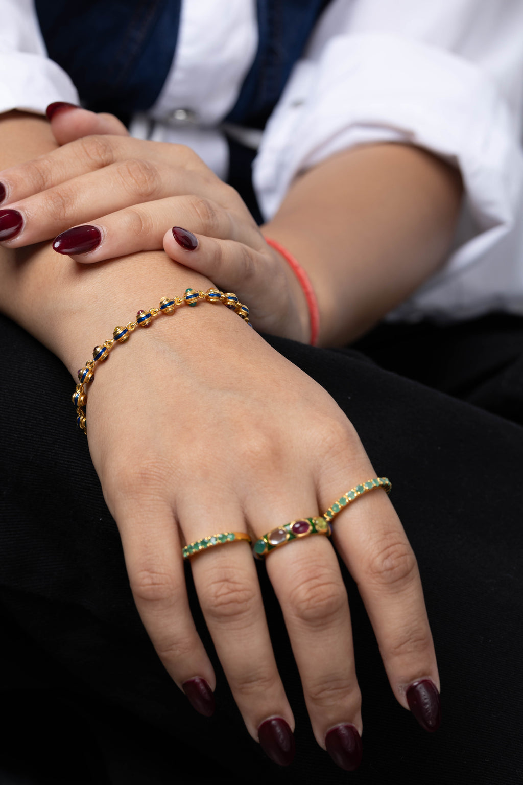 A close-up of a person's hands wearing a gold beaded bracelet and three matching gold rings with small green and red stones, paired with a white shirt and black pants.