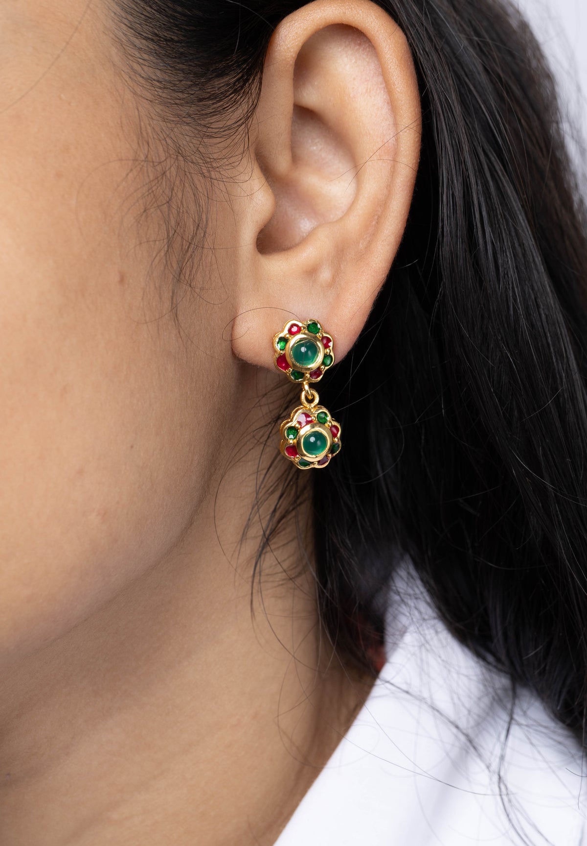 A close-up of a model's ear wearing a gold dangle earring with green and red enamel floral details.