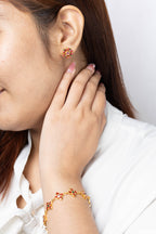 A close-up lifestyle shot of a woman wearing a gold-toned floral stud earring and a matching gold-toned floral bracelet, both accented with small red stones, against a clean white background.