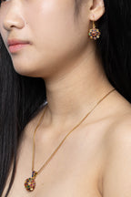 A close-up lifestyle shot of a model wearing a gold necklace with a colorful round pendant and a matching gold drop earring with a floral multicolor design.
