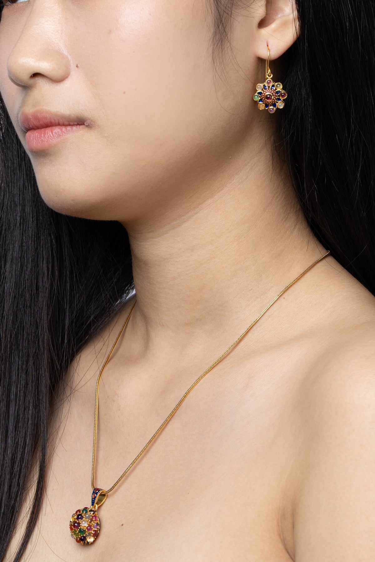 A close-up lifestyle shot of a model wearing a gold necklace with a colorful round pendant and a matching gold drop earring with a floral multicolor design.