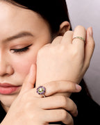 A close-up lifestyle shot of a woman resting her head on her hand, showcasing a silver ring with a purple and green gemstone cluster on her index finger, and another gold ring on her middle finger.