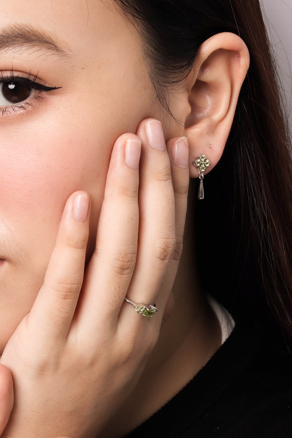 Close-up of a woman wearing a delicate gold ring with a small green gemstone cluster on her finger and a matching green gemstone dangle earring on her earlobe.