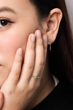 Close-up of a woman wearing a delicate gold ring with a small green gemstone cluster on her finger and a matching green gemstone dangle earring on her earlobe.
