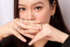 A close-up lifestyle shot of a woman wearing multiple silver rings with green and purple gemstones, holding her hands near her face against a light gray background.