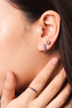 A close-up of a model's ear wearing a small floral stud earring and a vertical huggie hoop earring, with a matching ring on her finger.