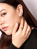 Close-up of a woman wearing delicate gold rings with small blue and pink gemstones, along with a matching earring, shown against a soft gray background.