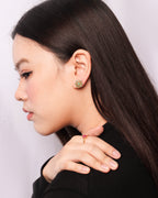 A woman in profile wearing a small green and gold floral stud earring and a delicate gold ring with a red gemstone on her finger, set against a plain light gray background.