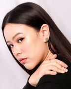A close-up portrait of a woman with long dark hair, wearing a small green floral stud earring and a gold ring with a red gemstone.