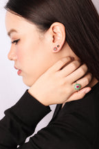 A close-up side profile of a woman wearing a gold ring with a red gemstone and green accents, along with matching stud earrings, against a light gray background.