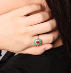 Close-up side profile of a woman wearing a small green and red stud earring and a matching green ring with a red center on her finger, against a light gray background.