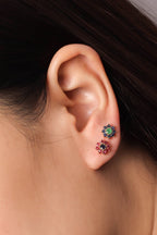 Close-up of a person's ear wearing two small flower-shaped stud earrings, one with green stones and one with red stones.