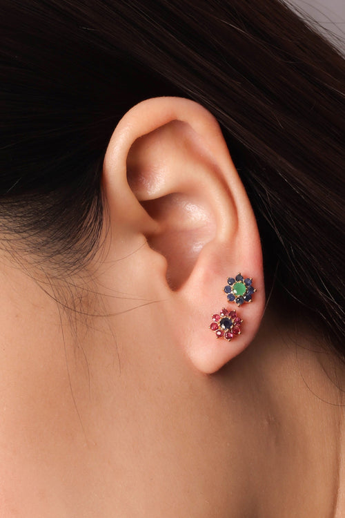 Close-up of a person's ear wearing two small flower-shaped stud earrings, one with green stones and one with red stones.